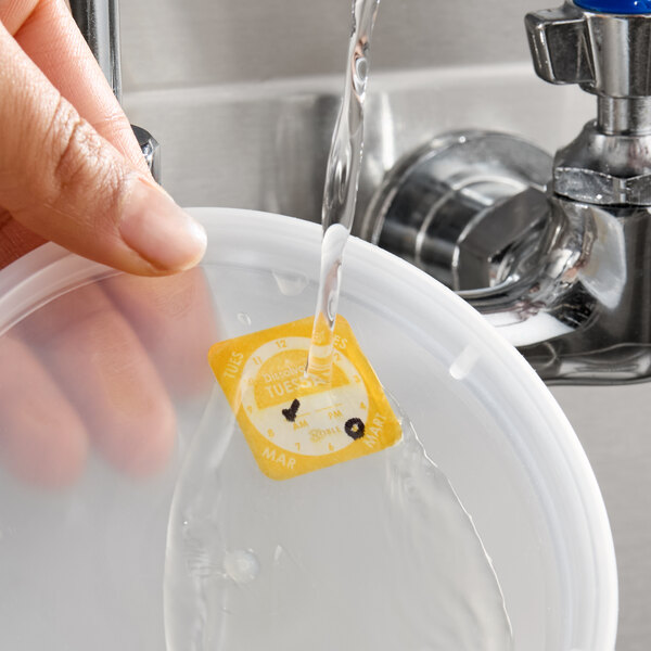 A dissolvable day of the week clock label being rinsed off a plastic container lid under running water.