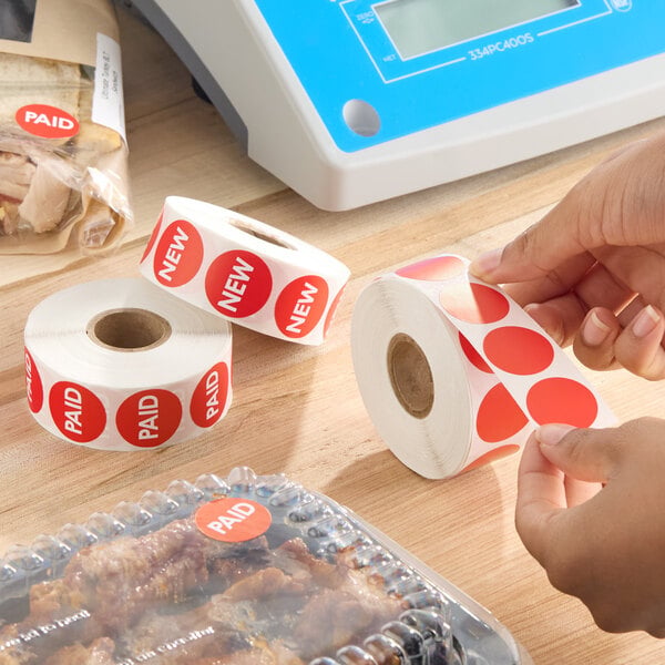 A roll of 1-inch round red blank labels being peeled and used, with other rolls labeled 'PAID' and 'NEW' nearby.
