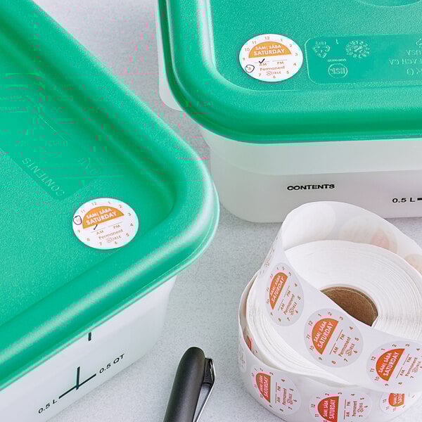 A green plastic container with a roll of Noble Products Saturday day of the week stickers on a table.