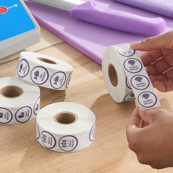 A roll of purple PointPlus fish allergen labels, each labeled as an allergen warning, with several similar rolls for other allergens on a wooden surface.