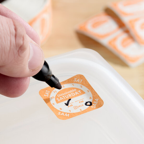 A dissolvable Saturday day of the week clock label being written on and applied to a food container.