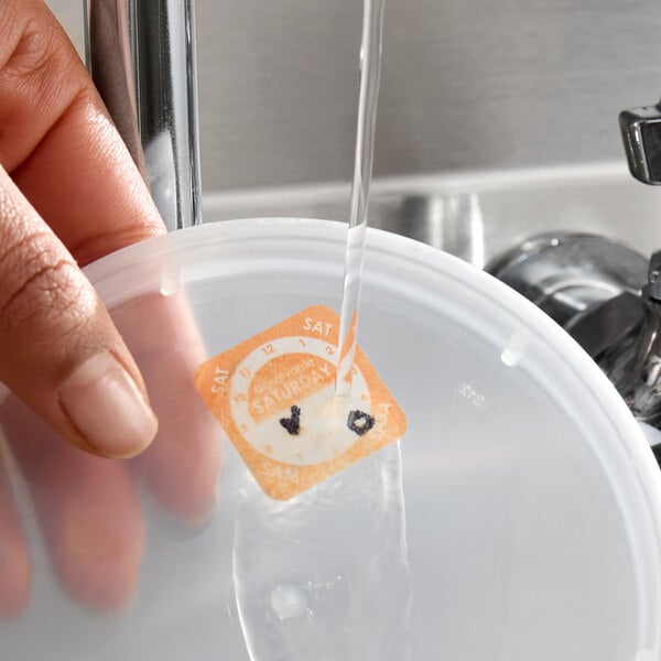 A dissolvable day of the week clock label marked for Saturday being rinsed off a plastic container lid under running water.