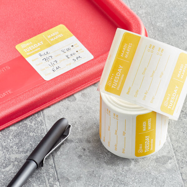 A roll of Noble Products Tuesday 2" x 2" Dissolvable Day of the Week labels next to a black pen with a red strap on a counter.