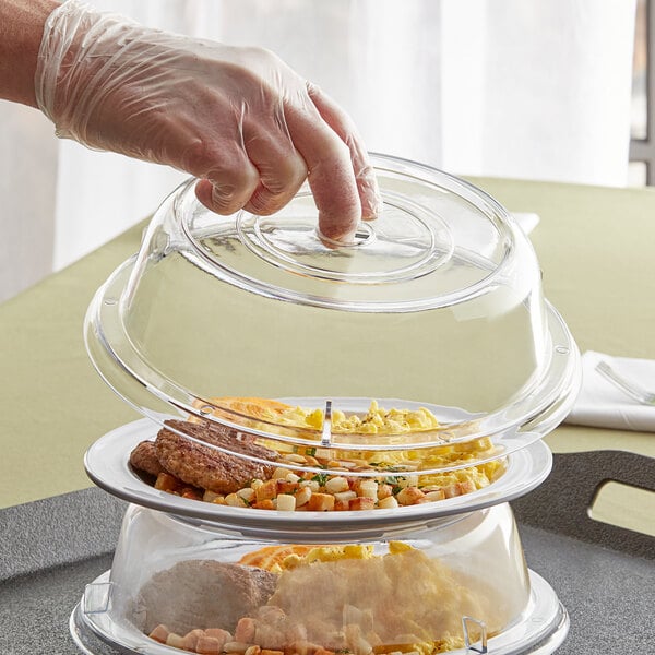 A hand in a plastic glove holding a clear polycarbonate Choice plate cover over a stack of food.