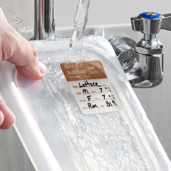 A dissolvable day of the week label marked 'Thursday' being washed off a food container under running water.