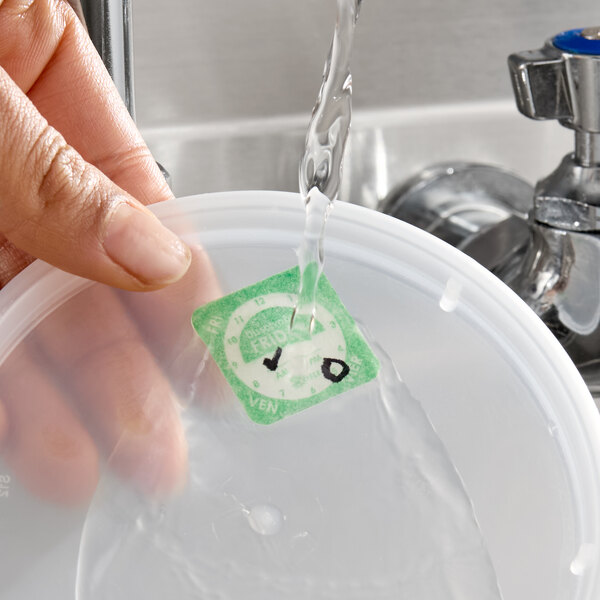 A dissolvable day of the week clock label being rinsed off a plastic container lid under running water.