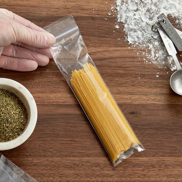 A hand holding a Clear Line plastic bag of spaghetti next to a bowl of dried herbs and a measuring spoon.