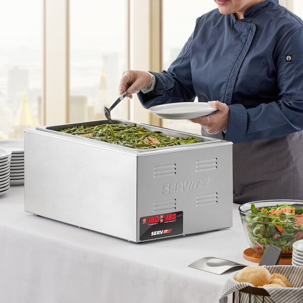 A woman in a chef's uniform using a ServIt countertop food warmer to serve food on a table.