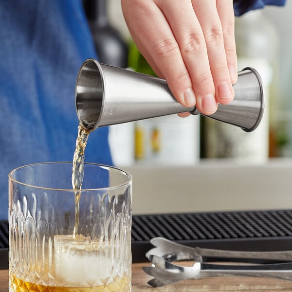 A hand using an Acopa stainless steel Japanese jigger to pour liquid into a glass on a metal bar.