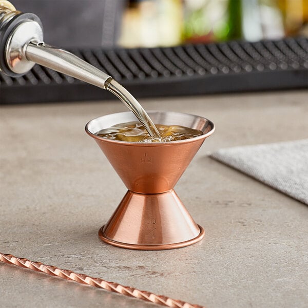 A copper jigger with liquid being poured into it using a metal nozzle.