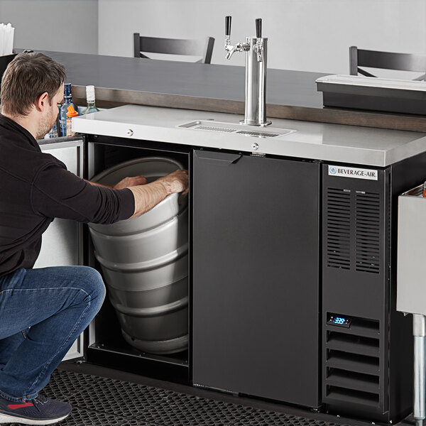 A man in a black shirt and jeans opening a keg in a black Beverage-Air beer cooler.