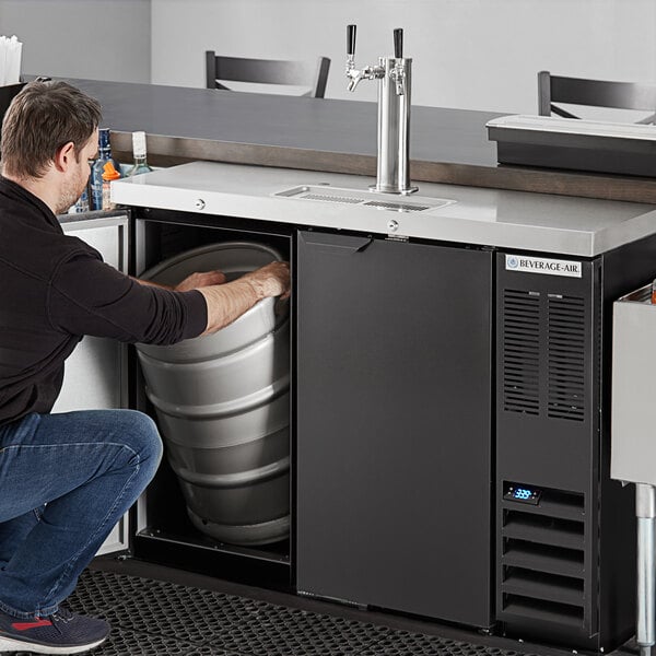 A man in a black shirt and jeans kneeling to open a silver keg in a black Beverage-Air beer cooler.