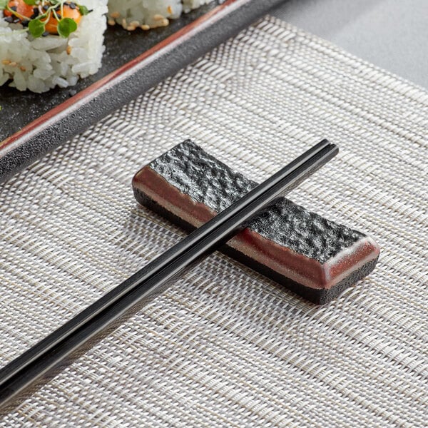 Acopa Heika black matte textured stoneware chopstick rest with chopsticks on a sushi plate.