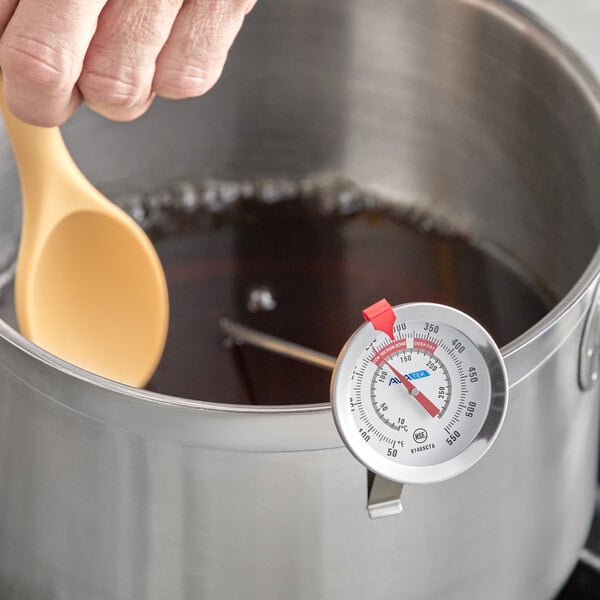 A stainless steel pot with a candy or deep fry probe thermometer attached to the rim and a hand stirring the contents with a yellow spoon.