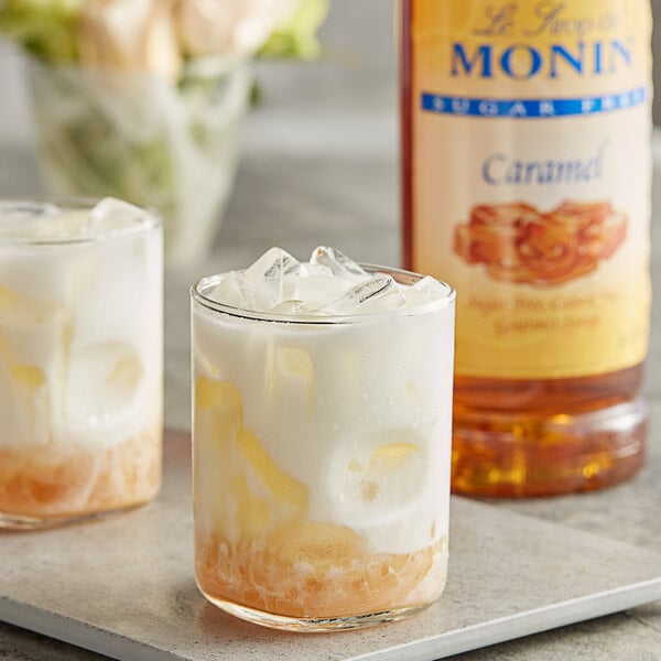 Two glasses of caramel flavored liquid and a bottle of Monin Sugar Free Caramel Flavoring Syrup.