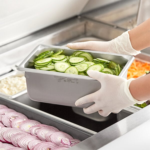 A person in gloves holding a Vollrath stainless steel steam table pan of cucumbers.