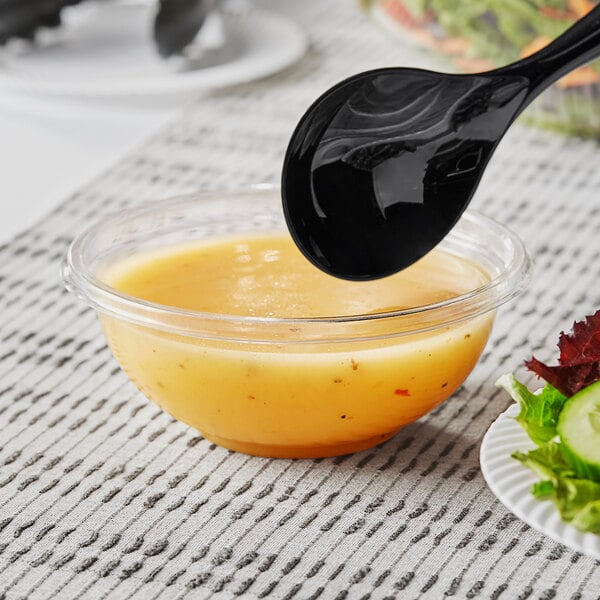 A clear Visions plastic bowl filled with salad, with a spoon and a jar of dressing.