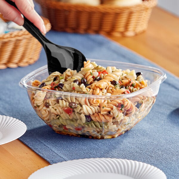 A hand holding a black spoon over a Visions clear plastic catering bowl of pasta.