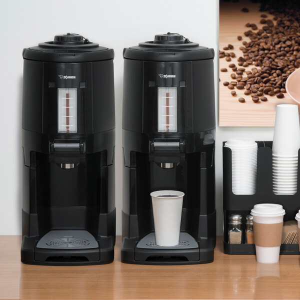 A Zojirushi stainless steel thermal gravity beverage dispenser on a table with a coffee machine and cups.
