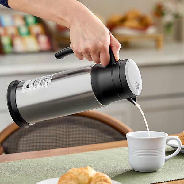 A hand pouring milk from a silver Zojirushi dairy server into a cup.