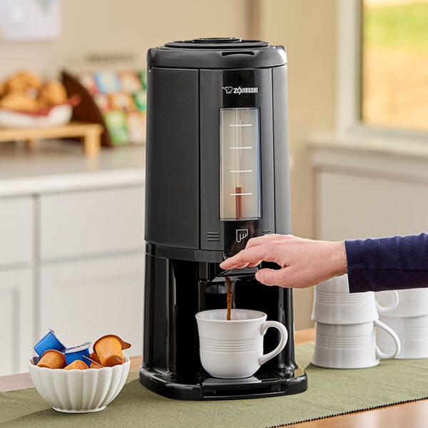A person using a Zojirushi glass-lined beverage dispenser to pour coffee into a white mug.