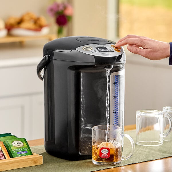 A person using a Zojirushi water dispenser to fill a glass mug with tea.