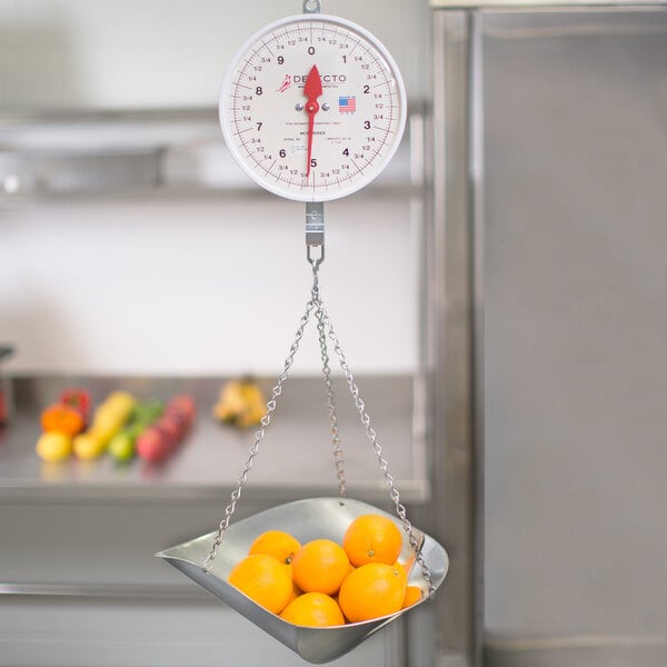A Cardinal Detecto hanging scoop scale with oranges in a metal scoop.