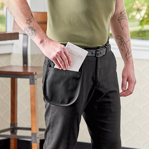 A man in a professional kitchen putting a piece of paper in a black Uncommon Chef utility pouch apron pocket.