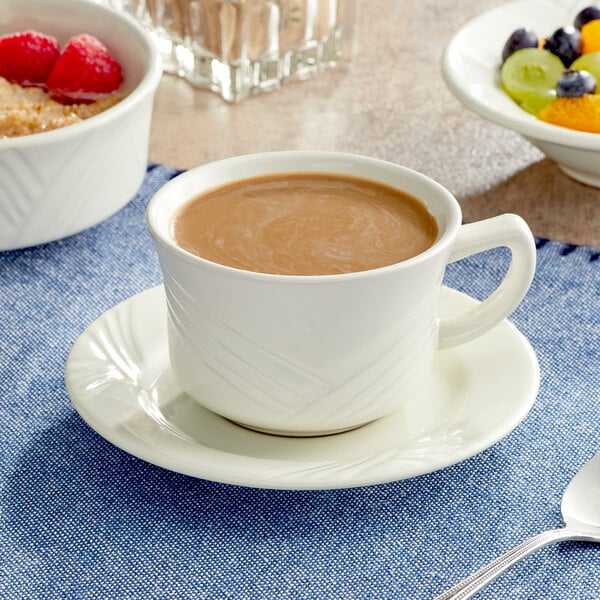 An Acopa Swell ivory stoneware cup filled with coffee sitting on a blue cloth next to a bowl of fruit.