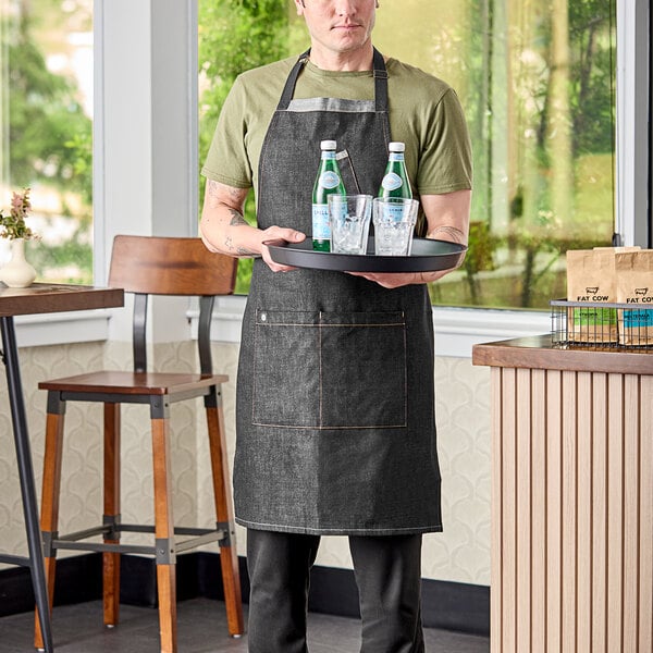 A man wearing a black Uncommon Chef denim apron holding a tray with drinks.