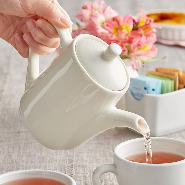 A person pouring water into a Tuxton eggshell white teapot.