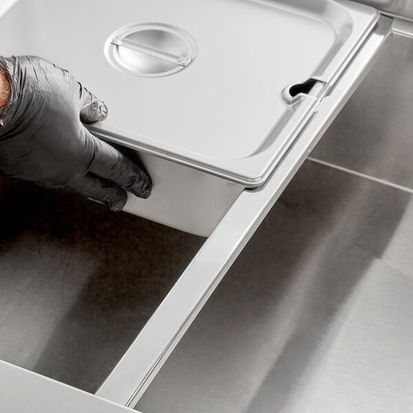 A person wearing black gloves using an Avantco stainless steel hotel pan adapter bar to hold a metal container.