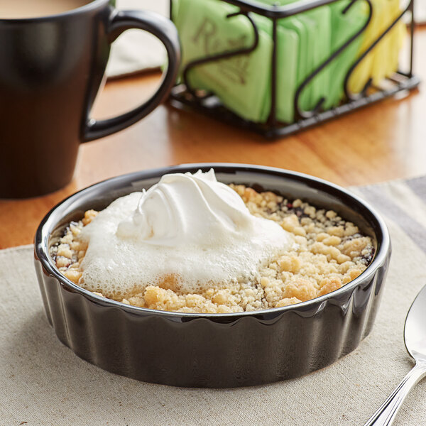 A glossy black fluted Acopa souffle dish filled with a dessert and whipped cream with a spoon on a table.