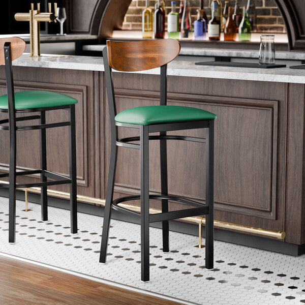 A bar stool with a black metal frame, green vinyl seat, and a curved vintage wood backrest.