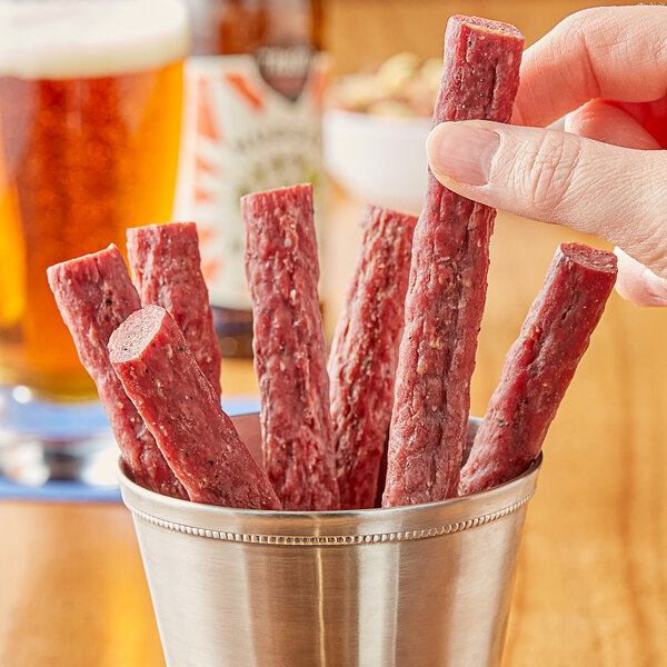 A person holding a silver cup filled with Kinikin Processing Rocky Mountain peppered elk snack sticks.