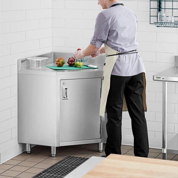 A person in a white shirt and apron cutting vegetables on a Regency stainless steel corner work table with a hinged door.