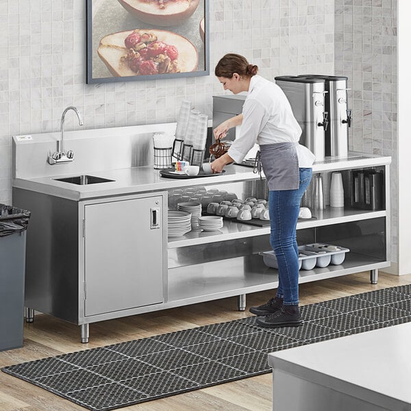 A woman standing in a school kitchen preparing food on a stainless steel Regency beverage table with a sink.