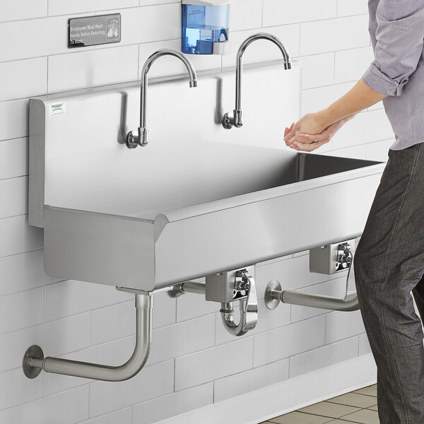 A person washing their hands in a wall mounted Regency multi-station hand sink with knee operated faucets.
