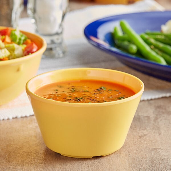 A yellow bowl of Acopa Foundations yellow melamine bouillon filled with soup on a table with a blue plate of green beans.