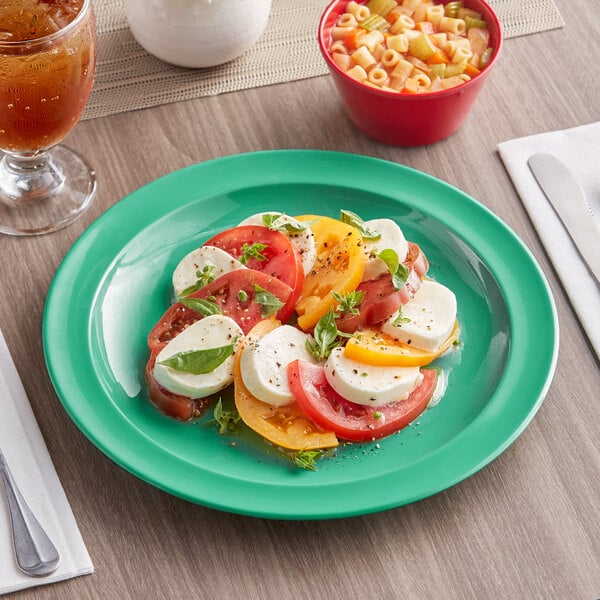 A green Acopa Foundations melamine plate holding a bowl of pasta, tomatoes, and cheese on a table.