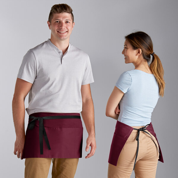 A man and woman wearing Choice burgundy waist aprons with black webbing standing in a professional kitchen.