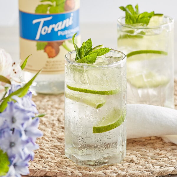 A glass of water with lime slices and mint leaves next to a Torani Puremade Zero Sugar Sweetener Syrup bottle.