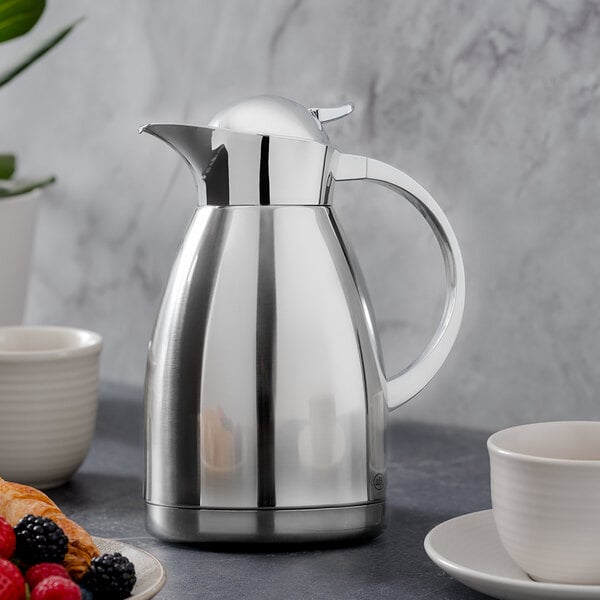 An Alfi stainless steel coffee carafe on a table with a cup of coffee.