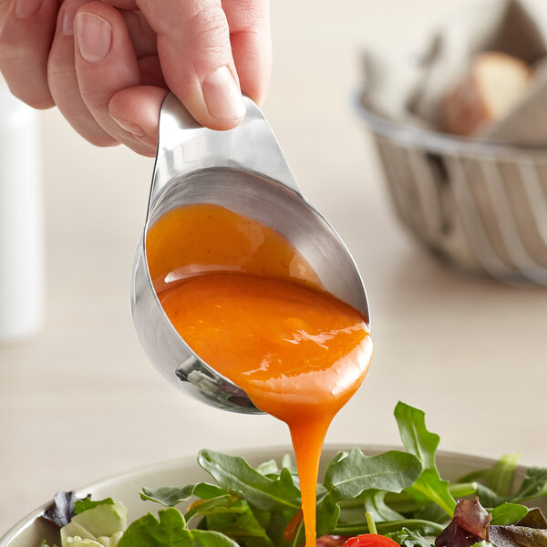 A person pouring orange sauce over a bowl of salad using a Vollrath stainless steel ramekin with a handle.