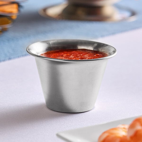 A Vollrath stainless steel round sauce cup with red sauce on a table.