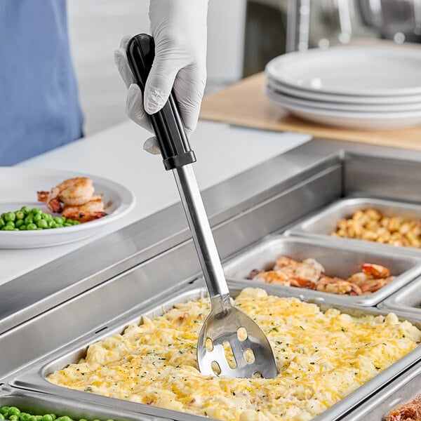 A hand holding an American Metalcraft stainless steel slotted spoon over food.