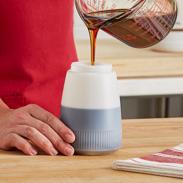 A person pouring brown syrup from a measuring cup into a white Vollrath syrup server.