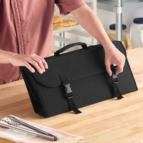 A person is handling a black carrying case with two buckle straps on a wooden countertop.