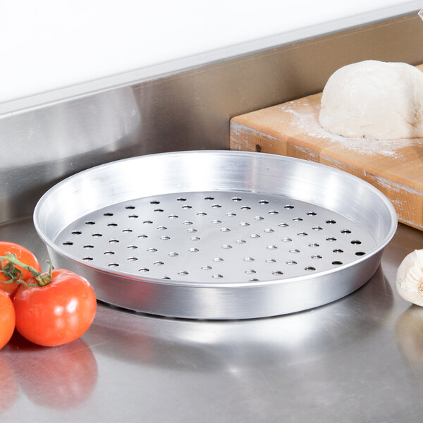 An American Metalcraft silver aluminum pizza pan with holes on a metal surface with tomatoes and dough.