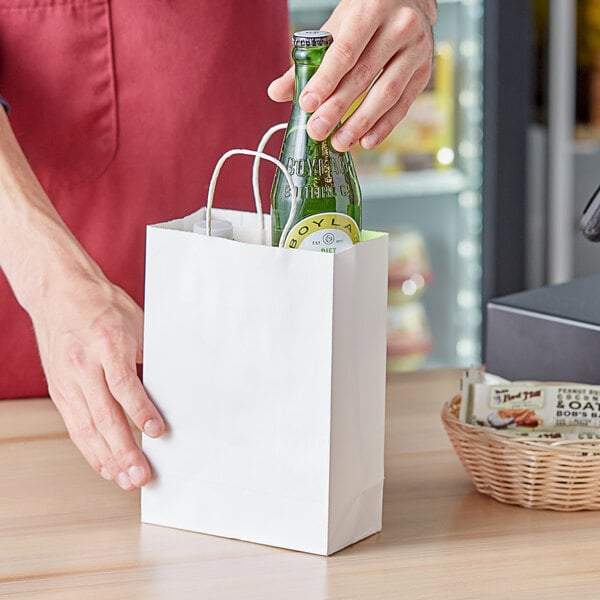 A person putting a green bottle into a white paper bag with handles.